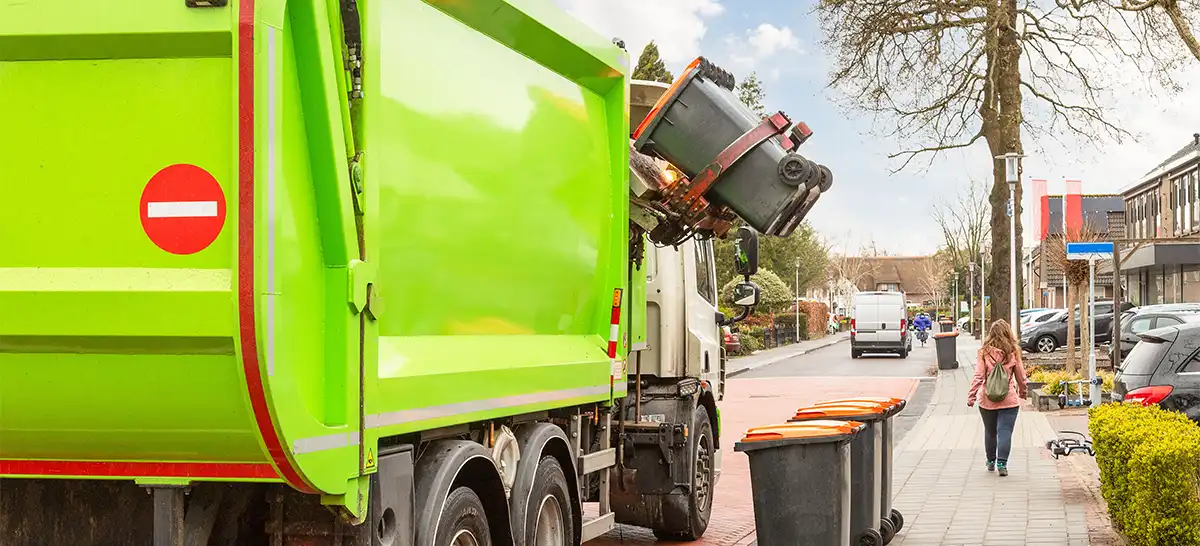 A refuse truck with an automated side loader body lifting a bin on a street.