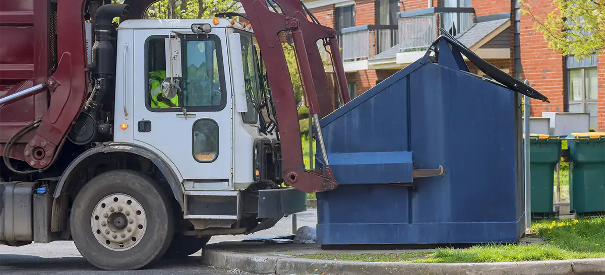 A front-loading refuse truck that is about to lift a large metal dumpster over its cab.